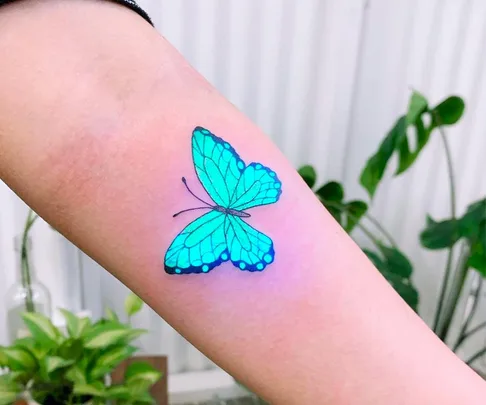 A glowing blue butterfly tattoo on a person's arm, set against a blurred plant background, showcasing UV ink.