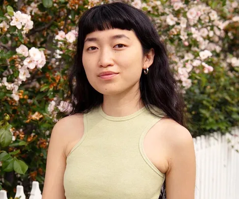 Woman with long dark hair and bangs standing in front of a white fence and pink flowering bushes, wearing a sleeveless top.