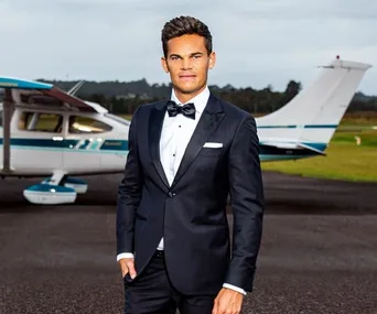 A man in a suit stands confidently in front of a small plane on the tarmac, with a cloudy sky and trees in the background.