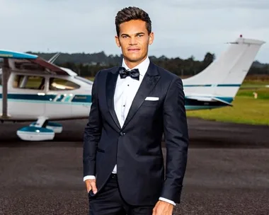 A man in a suit stands confidently in front of a small plane on the tarmac, with a cloudy sky and trees in the background.