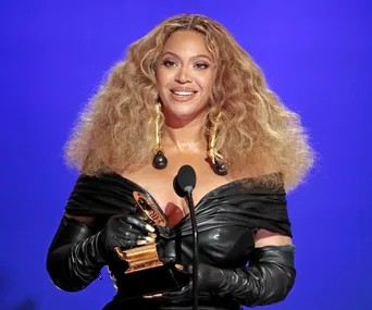 Beyoncé holding a Grammy award, wearing a black outfit, smiling on stage, blue background.