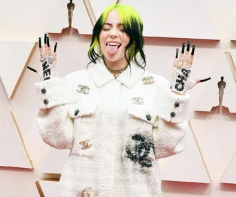 Person with black and green hair, white outfit, sticks out tongue, showing nails and jewelry on a red carpet event.