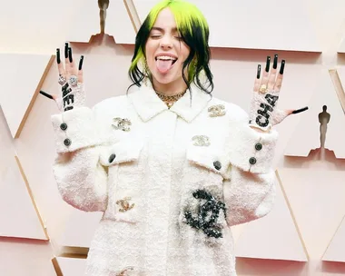 Person with black and green hair, white outfit, sticks out tongue, showing nails and jewelry on a red carpet event.