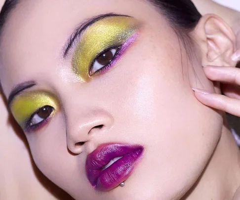 Close-up of a model wearing vibrant green and purple eye makeup with bold purple lipstick, showcasing Byredo cosmetics.