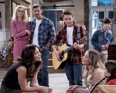 A group of people gathered in a living room, with one person playing a guitar, from the Netflix show "Country Comfort."