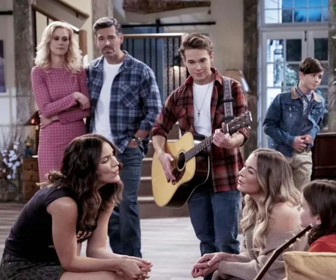 A group of people gathered in a living room, with one person playing a guitar, from the Netflix show "Country Comfort."