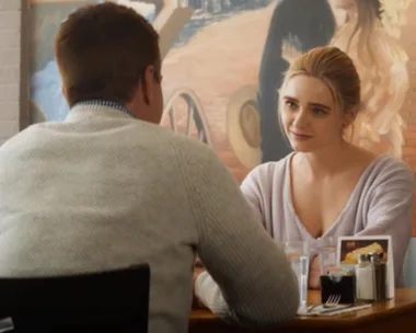 A woman and man sit across from each other at a table in a restaurant, with a painting in the background.