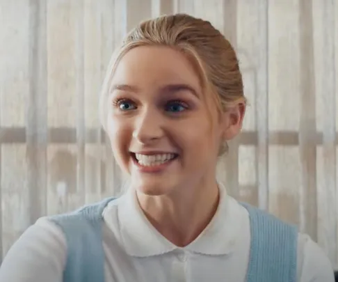 Young woman smiling brightly, wearing a white blouse and light blue vest, standing in front of a curtain.