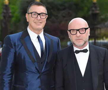 Two men in formal suits and glasses outdoors, one in a blue suit, the other in a black tuxedo.