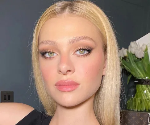 Blonde woman with flawless makeup and long lashes, posing indoors with tulips in the background.