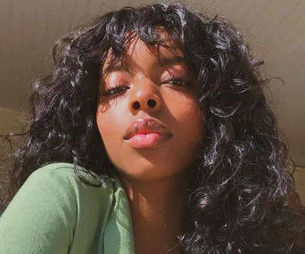 A woman with curly hair and bangs looks into the camera, wearing a green top. Bright natural lighting highlights her features.