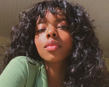 A woman with curly hair and bangs looks into the camera, wearing a green top. Bright natural lighting highlights her features.