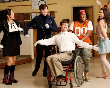 Cast of "Glee" performing energetically in a classroom setting; features a character in a wheelchair smiling.