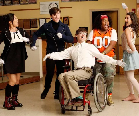 Cast of "Glee" performing energetically in a classroom setting; features a character in a wheelchair smiling.