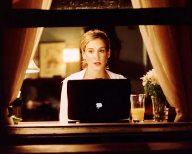 A person sitting at a window, working on a laptop, with a glass of juice and flowers nearby.