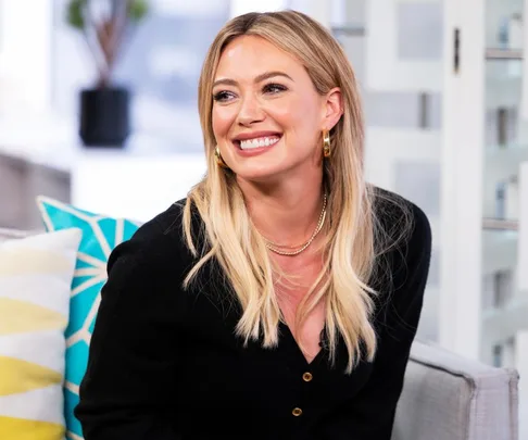 Smiling woman with blonde hair and gold earrings, sitting on a couch with patterned pillows.