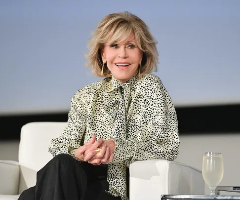 A person with short, wavy hair wearing a spotted blouse, sitting on a white chair, smiling with hands clasped.