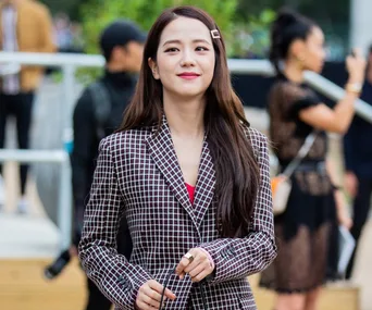 A person in a checkered blazer smiles slightly outdoors, with people blurred in the background.