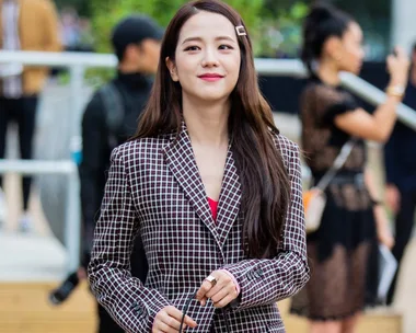 A person in a checkered blazer smiles slightly outdoors, with people blurred in the background.