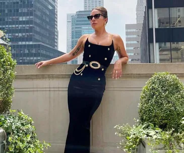 Woman in a sleek black dress and sunglasses poses on a balcony with city buildings in the background.