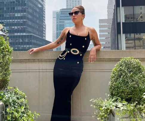 Woman in a sleek black dress and sunglasses poses on a balcony with city buildings in the background.