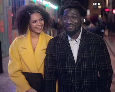 A woman in a bright coat and a man in a plaid jacket walk happily on a colorful, lively street at night.