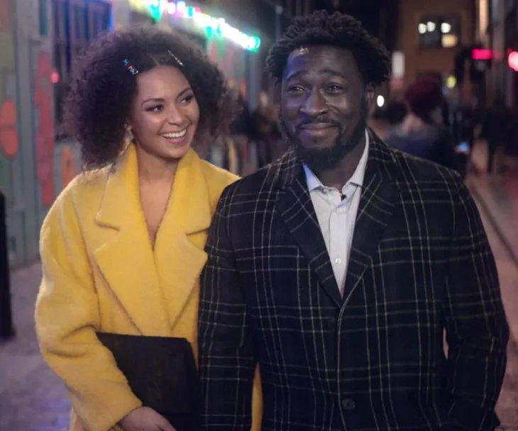 A woman in a bright coat and a man in a plaid jacket walk happily on a colorful, lively street at night.