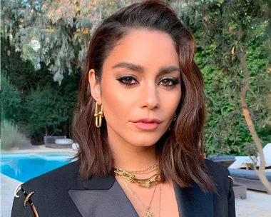 Woman with short brown hair and gold jewelry, wearing a black blazer, standing outdoors near a pool and greenery.