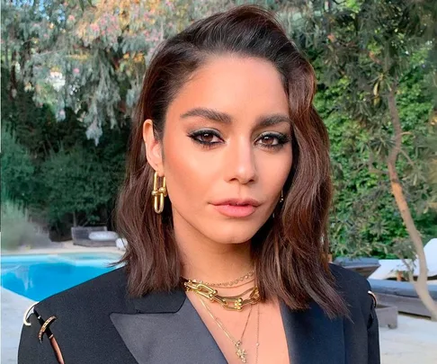 Woman with short brown hair and gold jewelry, wearing a black blazer, standing outdoors near a pool and greenery.