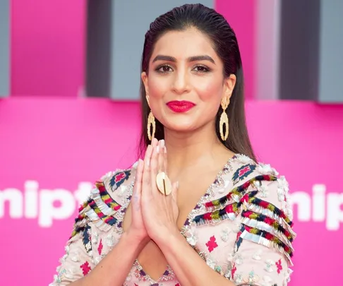 Woman in a colorful embroidered dress, smiling with hands in Namaste gesture against a pink background.