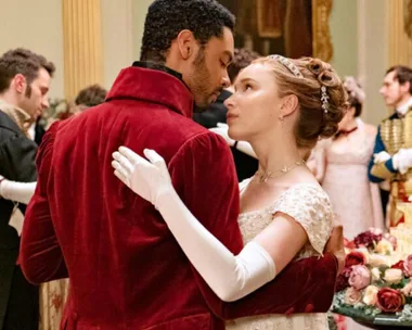 A couple in period attire dance intimately at a ballroom event, surrounded by elegantly dressed guests.