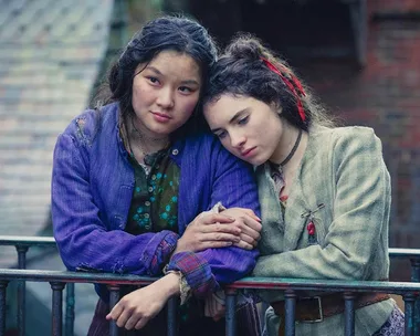 Two girls from "The Irregulars" lean on a railing, one in a purple jacket, both looking somber.