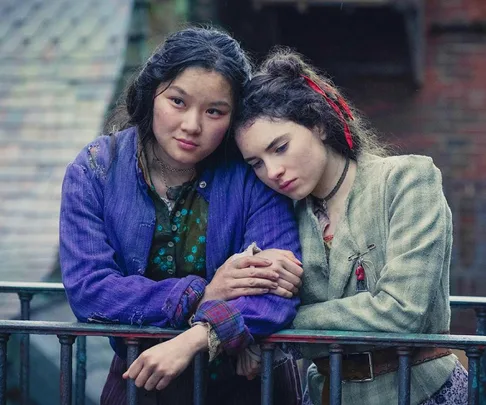 Two girls from "The Irregulars" lean on a railing, one in a purple jacket, both looking somber.