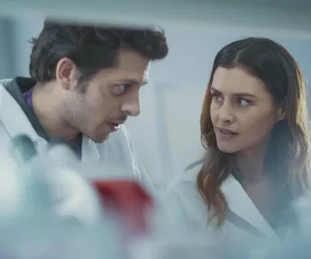 Two scientists wearing lab coats share a serious conversation in a laboratory setting.