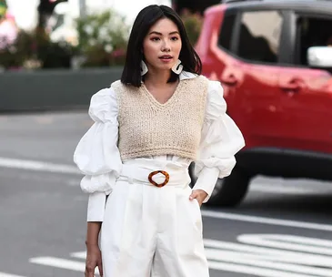 Woman in white blouse with puffy sleeves, beige knit vest, and white high-waisted trousers on city street.