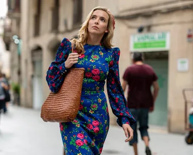 A woman wearing a blue dress with red flowers, carrying a brown woven bag, walking on a city street.