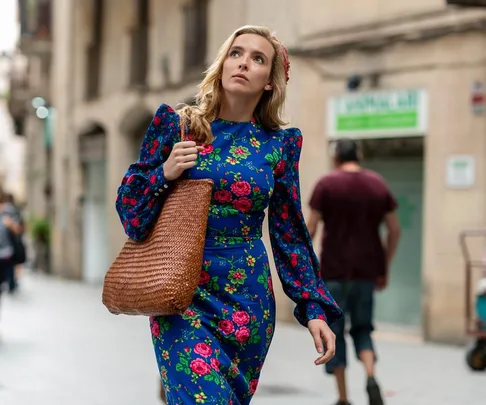 A woman wearing a blue dress with red flowers, carrying a brown woven bag, walking on a city street.