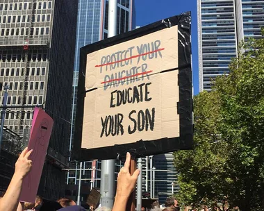 Protest sign reading "Protect your daughter" crossed out, replaced with "Educate your son" against city backdrop.