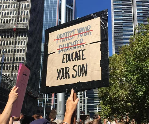 Protest sign reading "Protect your daughter" crossed out, replaced with "Educate your son" against city backdrop.