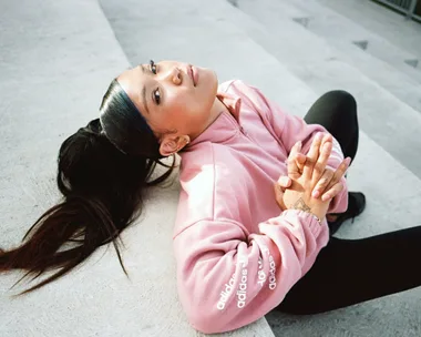 Woman in a pink Adidas jacket with a ponytail, sitting on steps with hands clasped.