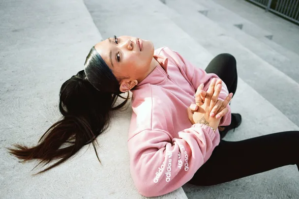 Woman in a pink Adidas jacket with a ponytail, sitting on steps with hands clasped.