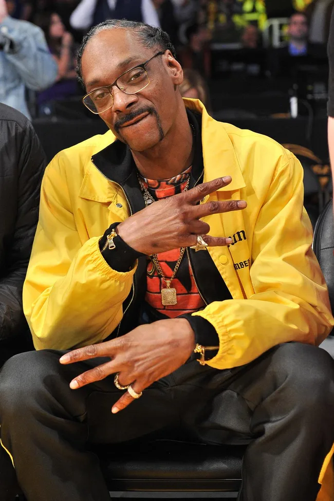 A person in a yellow jacket sits, making a peace sign with both hands, wearing glasses and jewelry.