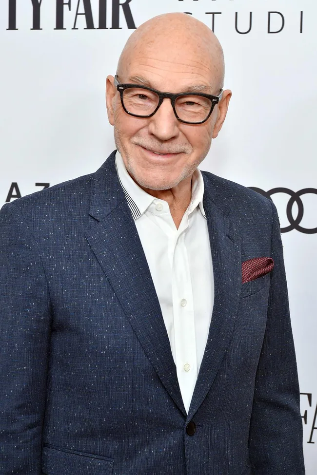 Man in a blue suit with glasses smiles at event, standing in front of a logo backdrop.