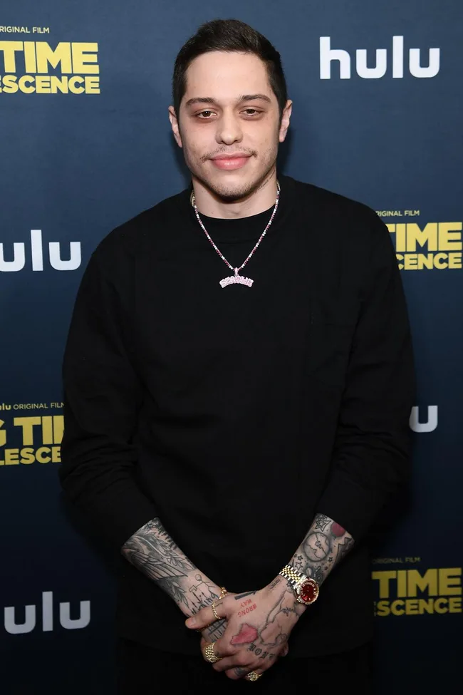 A person with tattoos wearing a black shirt and a necklace stands in front of a “Big Time Adolescence” Hulu backdrop.
