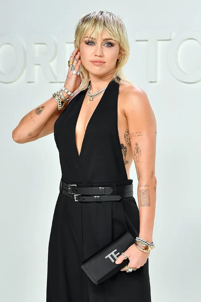 Person in a black dress with tattoos and jewelry, holding a black clutch with "TF" logo at a fashion event.