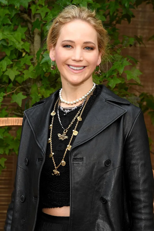 A woman in a black leather jacket and layered necklaces, smiling with green leaves in the background.