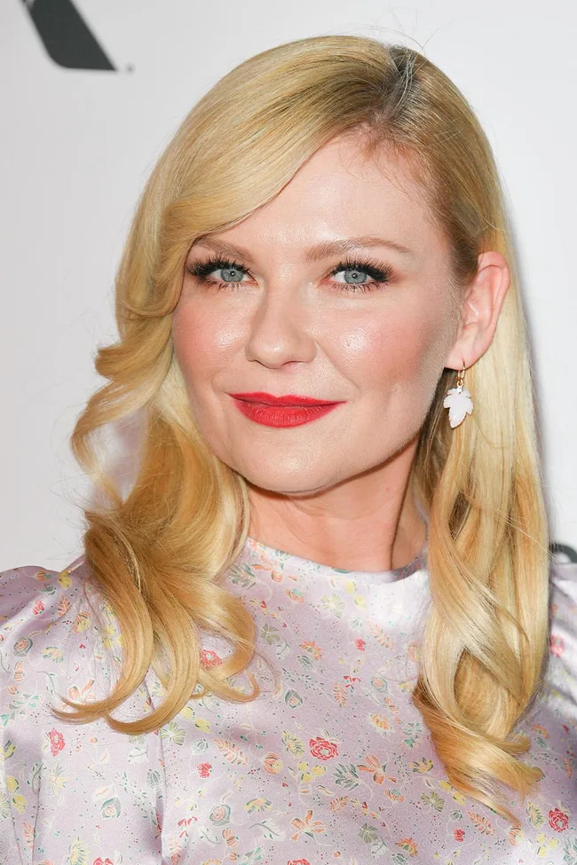A woman with long blonde hair, wearing a floral dress and red lipstick, smiles at the camera.