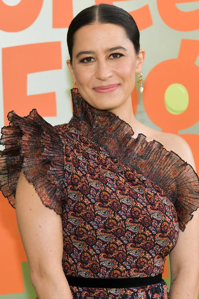 A woman in a patterned, asymmetrical dress smiles against a colorful background.