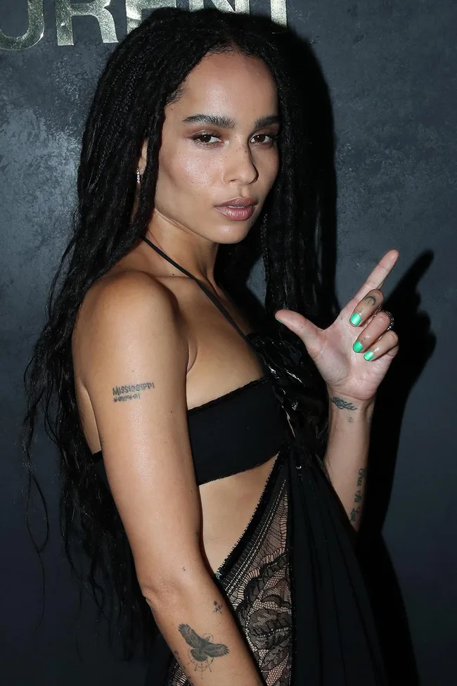 Woman with long hair and tattoos poses in a black dress, making a playful hand gesture.
