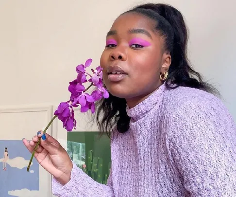 Woman in purple sweater holds a purple flower, with bold pink eye makeup, posing confidently indoors.